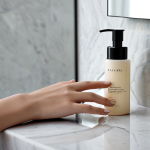A serene and elegant display of minimalist luxury beauty products, featuring 3-5 high-quality, sleek bottles and jars (e.g., a serum, a moisturizer, a cleanser) arranged neatly on a smooth, light-colored stone vanity. A female hand, adorned with modest, professional attire, gently hovers over one of the products, suggesting a thoughtful selection process. The background is a softly blurred modern bathroom with natural light, emphasizing cleanliness and sophistication. The overall mood is calm and refined, representing conscious beauty choices. Perfect anatomy, correct proportions, natural pose, well-formed hands, proper finger count, natural body proportions, fully clothed, modest clothing, appropriate attire, professional dress, safe for work, appropriate content, professional.